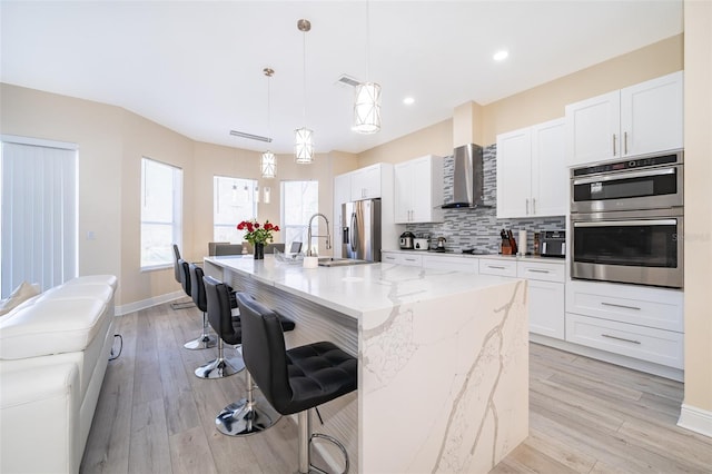 kitchen featuring decorative light fixtures, an island with sink, white cabinetry, stainless steel appliances, and wall chimney exhaust hood