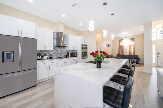 kitchen featuring wall chimney exhaust hood, white cabinetry, stainless steel fridge with ice dispenser, hanging light fixtures, and a kitchen island with sink