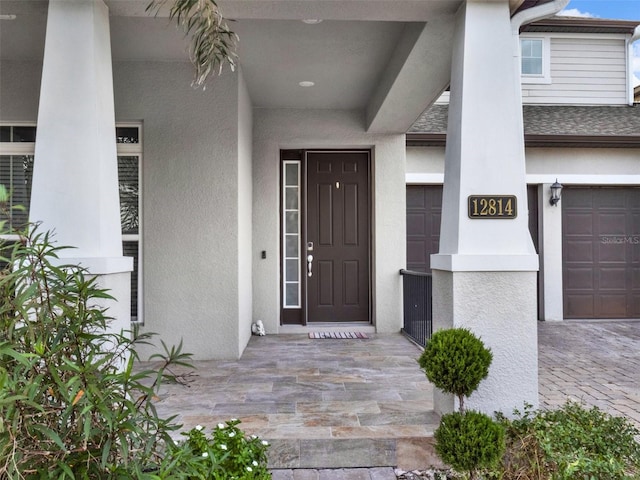 view of exterior entry featuring a garage, a shingled roof, decorative driveway, and stucco siding