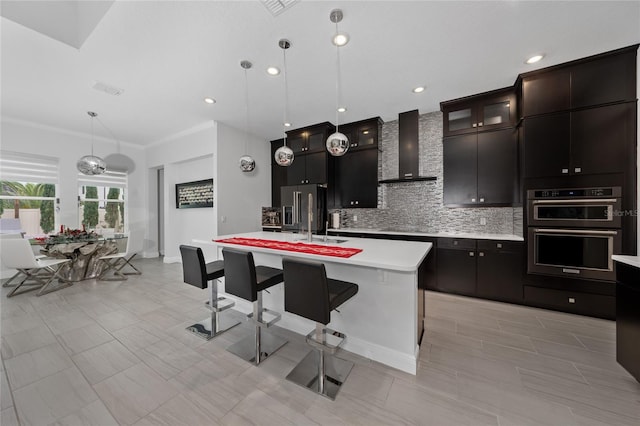 kitchen with wall chimney exhaust hood, tasteful backsplash, hanging light fixtures, appliances with stainless steel finishes, and a kitchen island with sink