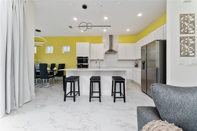 kitchen featuring wall chimney range hood, stainless steel appliances, an island with sink, and white cabinets