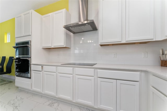 kitchen featuring wall chimney range hood, black electric stovetop, white cabinets, decorative backsplash, and stainless steel double oven