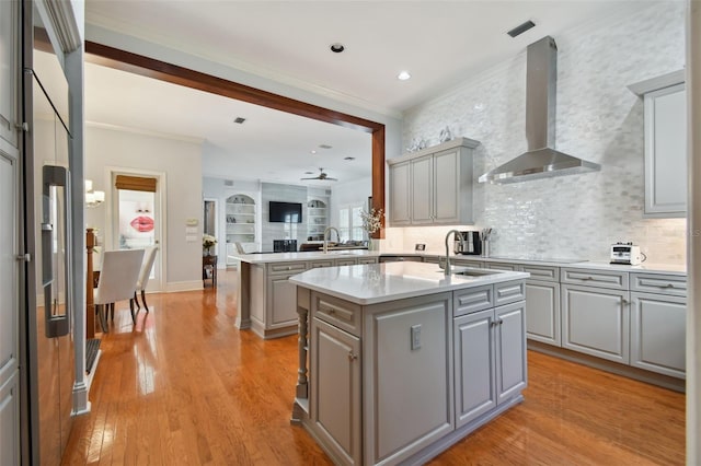 kitchen with wall chimney range hood, sink, a kitchen island with sink, gray cabinetry, and kitchen peninsula