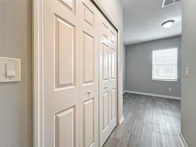 hall with a textured ceiling, a textured wall, wood finished floors, visible vents, and baseboards