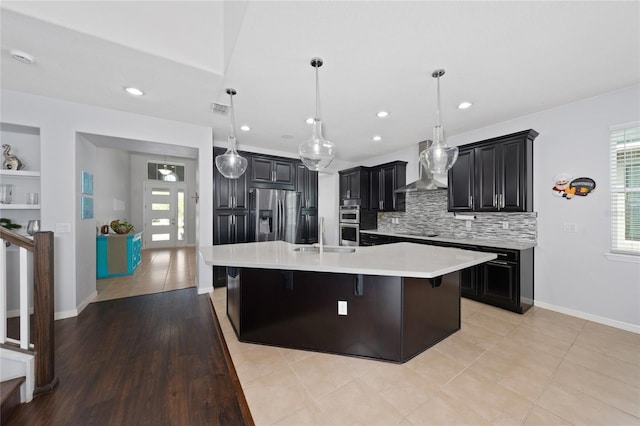 kitchen featuring a breakfast bar, an island with sink, hanging light fixtures, stainless steel appliances, and wall chimney exhaust hood