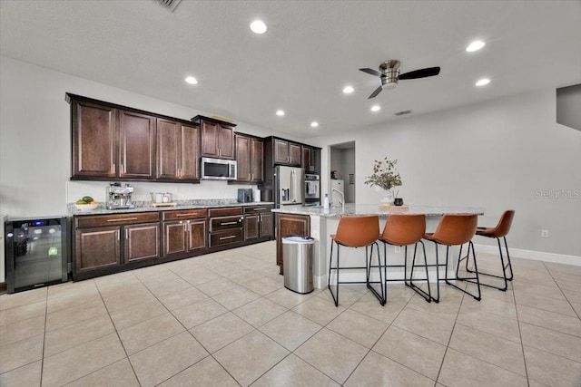 kitchen featuring light tile patterned flooring, a breakfast bar area, appliances with stainless steel finishes, an island with sink, and beverage cooler