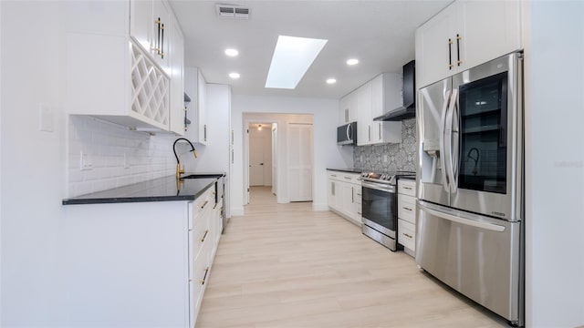 kitchen with appliances with stainless steel finishes, a skylight, sink, white cabinets, and wall chimney range hood