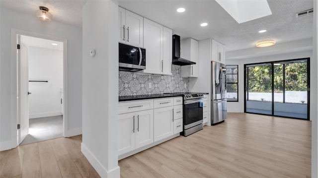 kitchen featuring a skylight, white cabinets, decorative backsplash, stainless steel appliances, and wall chimney range hood