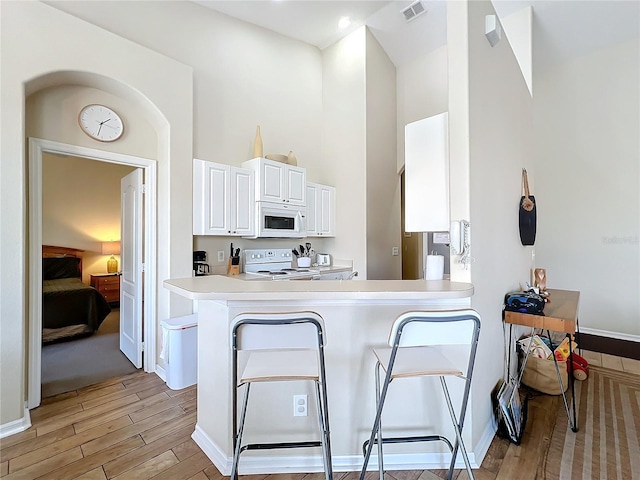 kitchen with a towering ceiling, white cabinets, kitchen peninsula, white appliances, and light hardwood / wood-style flooring