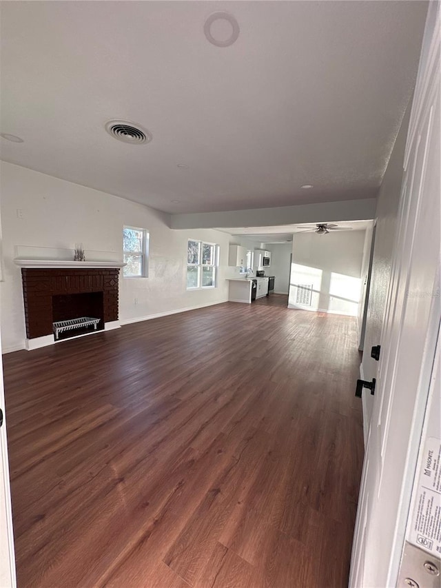 unfurnished living room with dark wood-style flooring, a brick fireplace, visible vents, and a ceiling fan