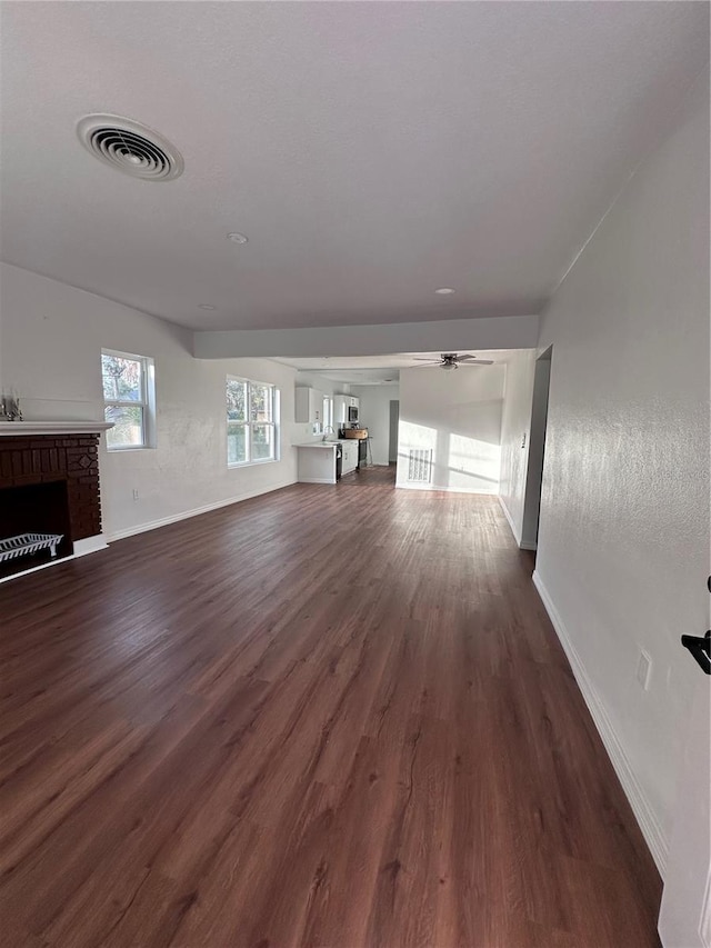 unfurnished living room with ceiling fan, a fireplace, visible vents, baseboards, and dark wood-style floors