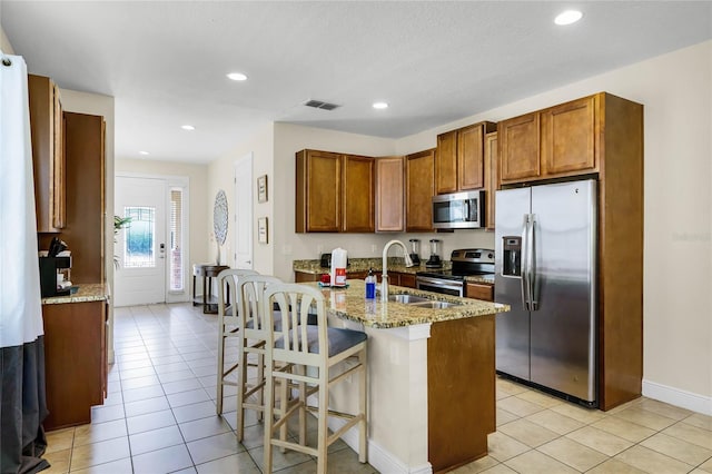 kitchen with light tile patterned floors, appliances with stainless steel finishes, a kitchen breakfast bar, light stone countertops, and a kitchen island with sink