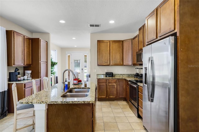 kitchen with sink, light stone counters, light tile patterned floors, appliances with stainless steel finishes, and an island with sink