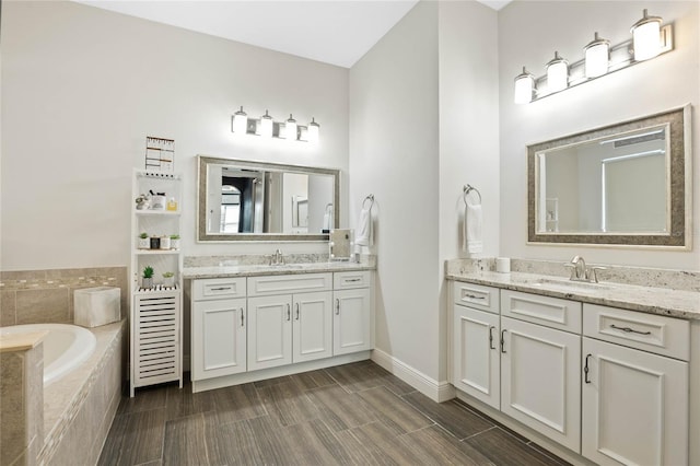 full bath featuring a relaxing tiled tub, two vanities, a sink, and baseboards