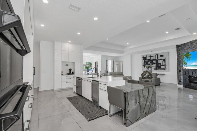 kitchen with a breakfast bar, white cabinetry, light stone counters, a center island with sink, and a raised ceiling