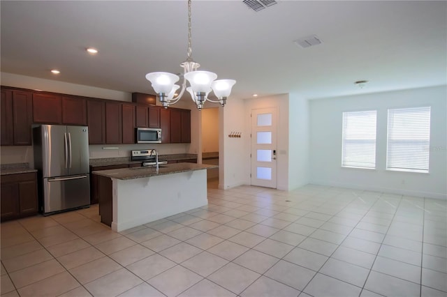 kitchen with decorative light fixtures, light tile patterned floors, dark stone countertops, appliances with stainless steel finishes, and an island with sink