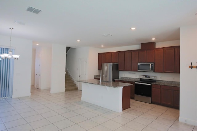 kitchen with light tile patterned floors, appliances with stainless steel finishes, dark stone countertops, hanging light fixtures, and a center island with sink