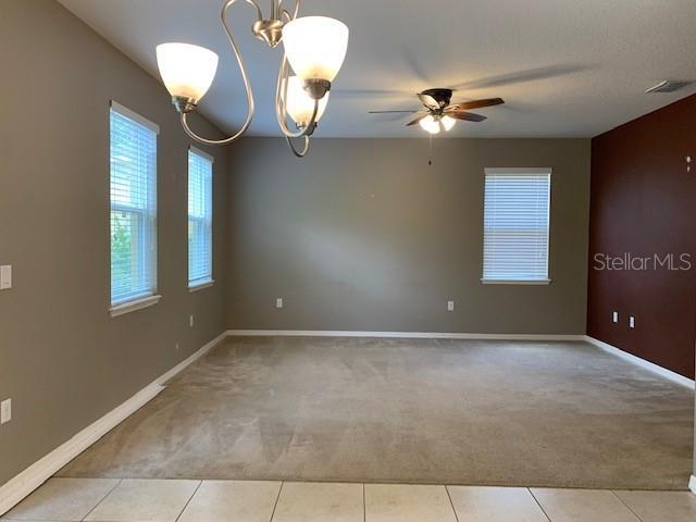 carpeted empty room featuring ceiling fan with notable chandelier, tile patterned floors, baseboards, and visible vents