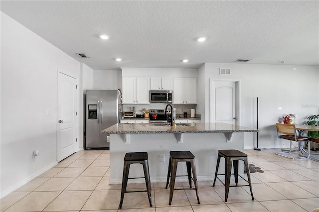 kitchen featuring white cabinets, dark stone counters, an island with sink, appliances with stainless steel finishes, and a kitchen bar