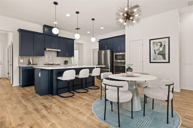 kitchen featuring a center island with sink, stainless steel appliances, light countertops, under cabinet range hood, and a kitchen breakfast bar