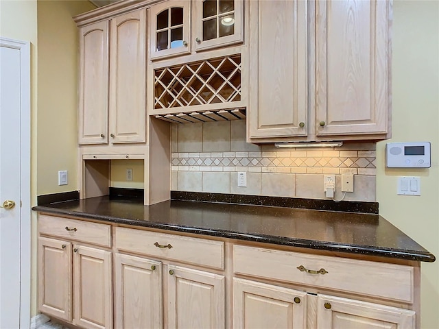 kitchen with light brown cabinets, glass insert cabinets, decorative backsplash, and dark stone countertops