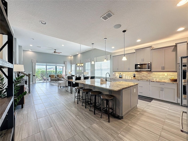 kitchen featuring gray cabinetry, visible vents, light stone countertops, stainless steel microwave, and an island with sink