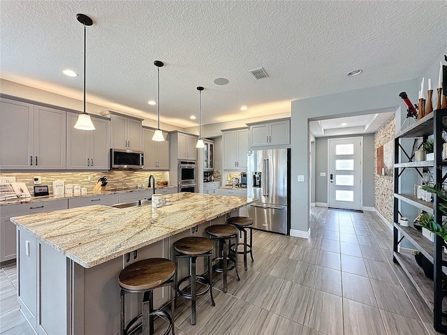 kitchen featuring stainless steel appliances, gray cabinets, a center island with sink, and hanging light fixtures