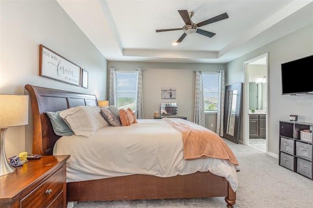 bedroom featuring light colored carpet, a ceiling fan, baseboards, a tray ceiling, and ensuite bath