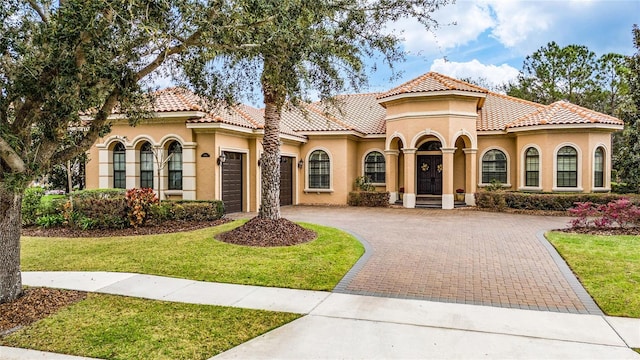 mediterranean / spanish house featuring an attached garage, a tile roof, decorative driveway, stucco siding, and a front yard