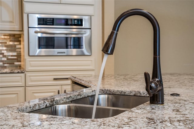 interior space with cream cabinetry, tasteful backsplash, a sink, light stone countertops, and oven