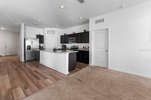 kitchen with light stone counters, a sink, visible vents, appliances with stainless steel finishes, and an island with sink