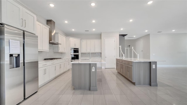 kitchen with an island with sink, backsplash, appliances with stainless steel finishes, wall chimney exhaust hood, and light countertops