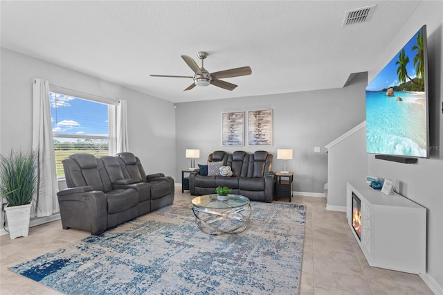 living area with ceiling fan, a textured ceiling, visible vents, baseboards, and a glass covered fireplace