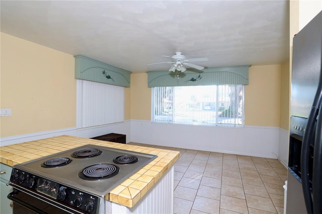 kitchen featuring light tile patterned floors, electric range oven, wainscoting, tile countertops, and black fridge with ice dispenser