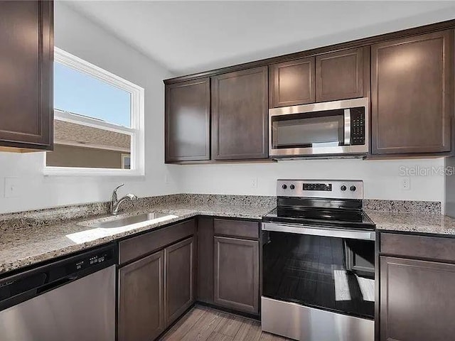 kitchen with stainless steel appliances, a sink, dark brown cabinets, light stone countertops, and light wood-type flooring