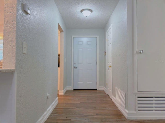 doorway with a textured ceiling, a textured wall, wood finished floors, visible vents, and baseboards