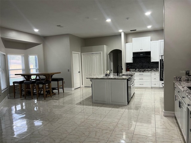 kitchen featuring marble finish floor, a kitchen island with sink, light stone counters, tasteful backsplash, and arched walkways