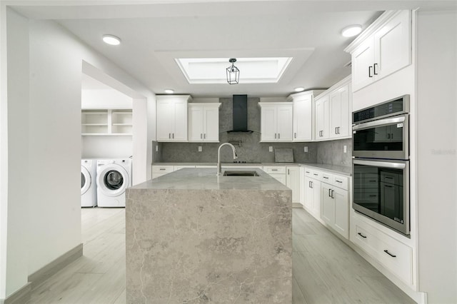 kitchen featuring tasteful backsplash, washing machine and clothes dryer, stainless steel double oven, wall chimney range hood, and white cabinetry