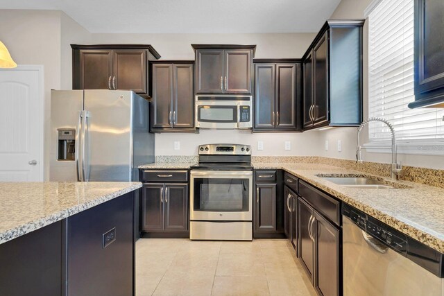 kitchen featuring light stone counters, light tile patterned floors, appliances with stainless steel finishes, dark brown cabinetry, and a sink