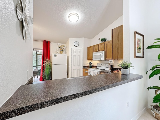 kitchen with a textured ceiling, a peninsula, white appliances, brown cabinetry, and dark countertops