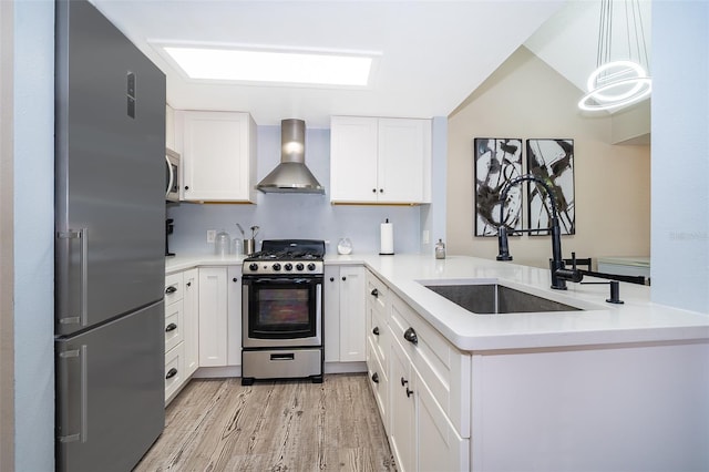 kitchen with light wood finished floors, stainless steel appliances, white cabinets, a sink, and wall chimney exhaust hood