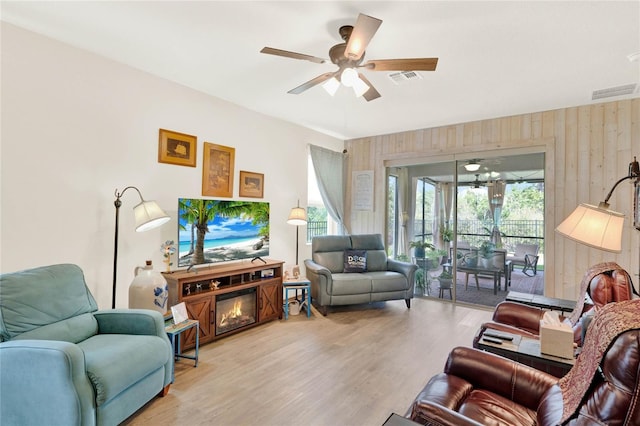 living room with wooden walls, wood finished floors, visible vents, ceiling fan, and a glass covered fireplace