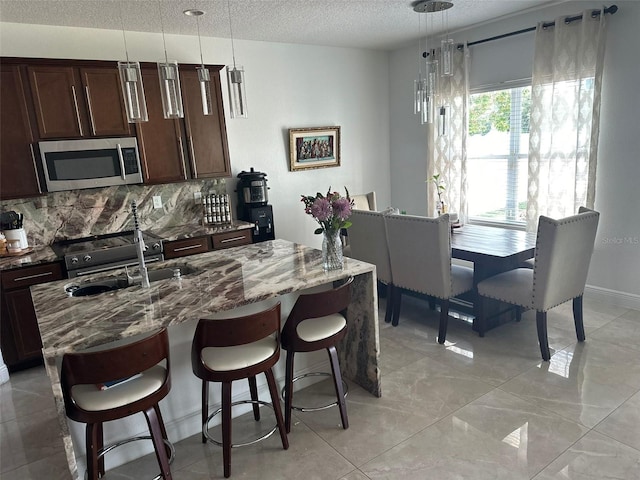 kitchen with a textured ceiling, stone counters, hanging light fixtures, decorative backsplash, and stainless steel microwave