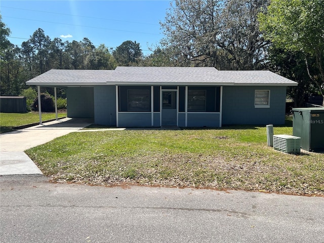 ranch-style house featuring concrete block siding, roof with shingles, a front yard, a carport, and driveway