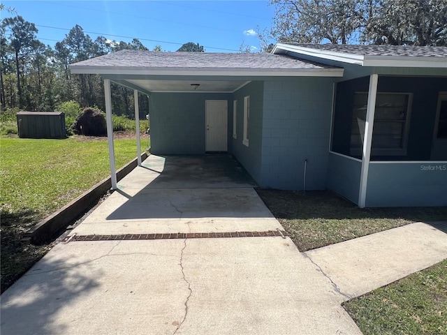 exterior space with a shingled roof, concrete block siding, concrete driveway, a lawn, and a carport