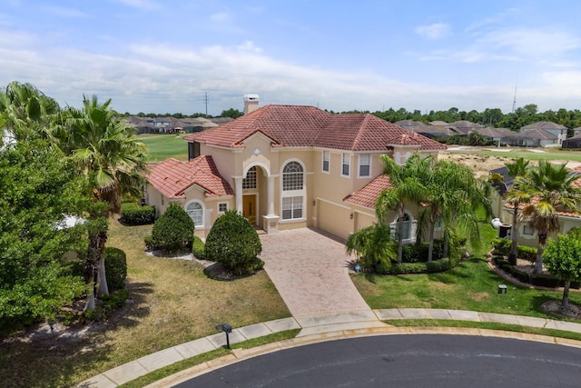 mediterranean / spanish house with a tiled roof, decorative driveway, stucco siding, a front lawn, and a chimney