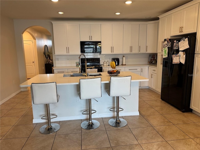 kitchen with black appliances, light tile patterned floors, arched walkways, and a kitchen island with sink