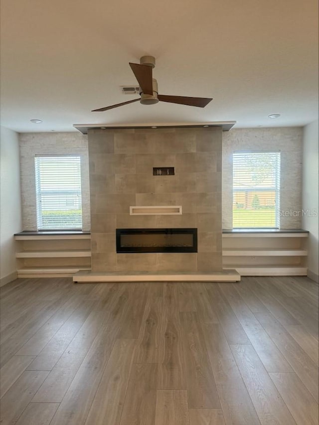 unfurnished living room with a tiled fireplace, plenty of natural light, ceiling fan, and wood finished floors