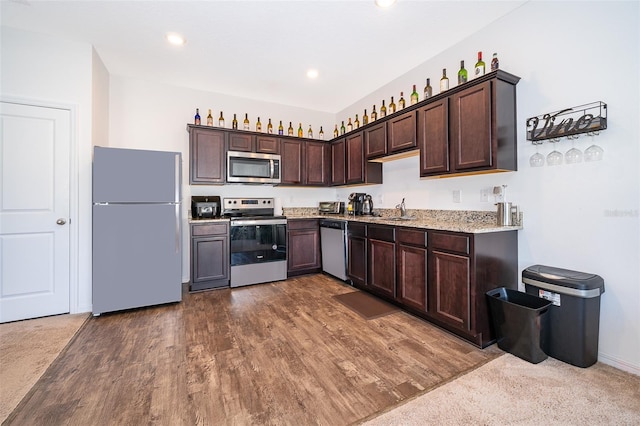 kitchen featuring light stone counters, dark brown cabinetry, stainless steel appliances, dark wood-style flooring, and a sink