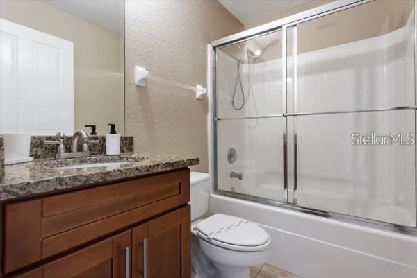 full bathroom with a textured wall, bath / shower combo with glass door, vanity, and toilet